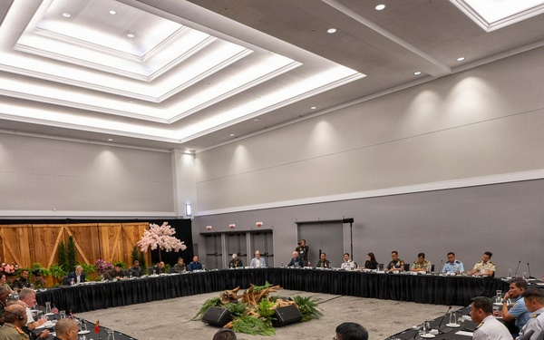 A large formal multilateral meeting or conference is taking place in a modern conference hall, with participants seated around a large square arrangement of tables with black skirting. The attendees appear to include military personnel in various uniforms and civilians, suggesting a high-level defense or diplomatic forum. The room is professionally decorated with floral arrangements and tropical plants in the center of the table configuration.