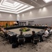 A large formal multilateral meeting or conference is taking place in a modern conference hall, with participants seated around a large square arrangement of tables with black skirting. The attendees appear to include military personnel in various uniforms and civilians, suggesting a high-level defense or diplomatic forum. The room is professionally decorated with floral arrangements and tropical plants in the center of the table configuration.