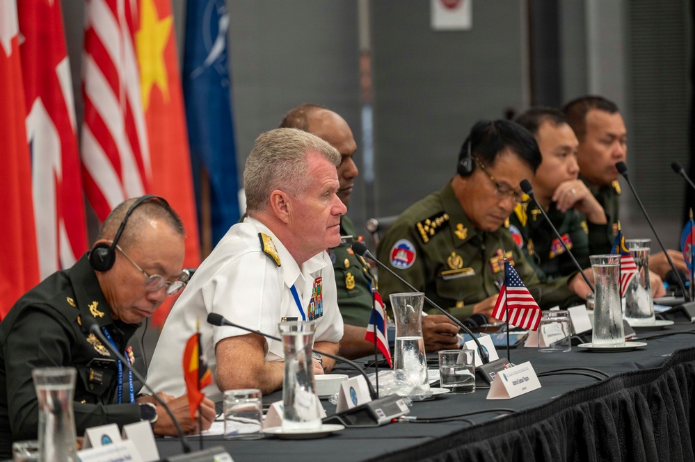 A multinational military conference is taking place with senior officers from multiple nations seated at a long table equipped with microphones and translation headsets. A US Navy admiral in white dress uniform appears to be speaking or presenting, flanked by military officers in various national uniforms including green combat dress. Small national flags, including a US flag, are visible on the table alongside name placards and water glasses.