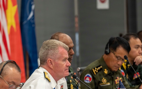 A multinational military conference is taking place with senior officers from multiple nations seated at a long table equipped with microphones and translation headsets. A US Navy admiral in white dress uniform appears to be speaking or presenting, flanked by military officers in various national uniforms including green combat dress. Small national flags, including a US flag, are visible on the table alongside name placards and water glasses.