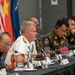 A multinational military conference is taking place with senior officers from multiple nations seated at a long table equipped with microphones and translation headsets. A US Navy admiral in white dress uniform appears to be speaking or presenting, flanked by military officers in various national uniforms including green combat dress. Small national flags, including a US flag, are visible on the table alongside name placards and water glasses.