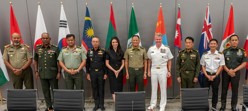 A formal group photograph of military officers and one civilian woman posing in front of multiple national flags. The gathering appears to be a multinational military meeting or conference, with personnel wearing uniforms from various nations. The setting is an indoor conference or meeting room with chairs visible in the foreground.