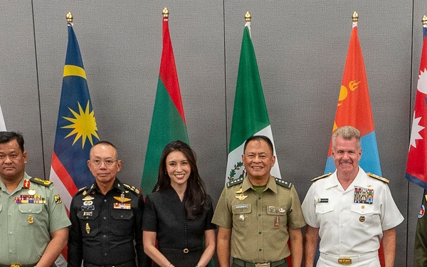 A formal group photograph of military officers and one civilian woman posing in front of multiple national flags. The gathering appears to be a multinational military meeting or conference, with personnel wearing uniforms from various nations. The setting is an indoor conference or meeting room with chairs visible in the foreground.