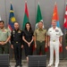 A formal group photograph of military officers and one civilian woman posing in front of multiple national flags. The gathering appears to be a multinational military meeting or conference, with personnel wearing uniforms from various nations. The setting is an indoor conference or meeting room with chairs visible in the foreground.