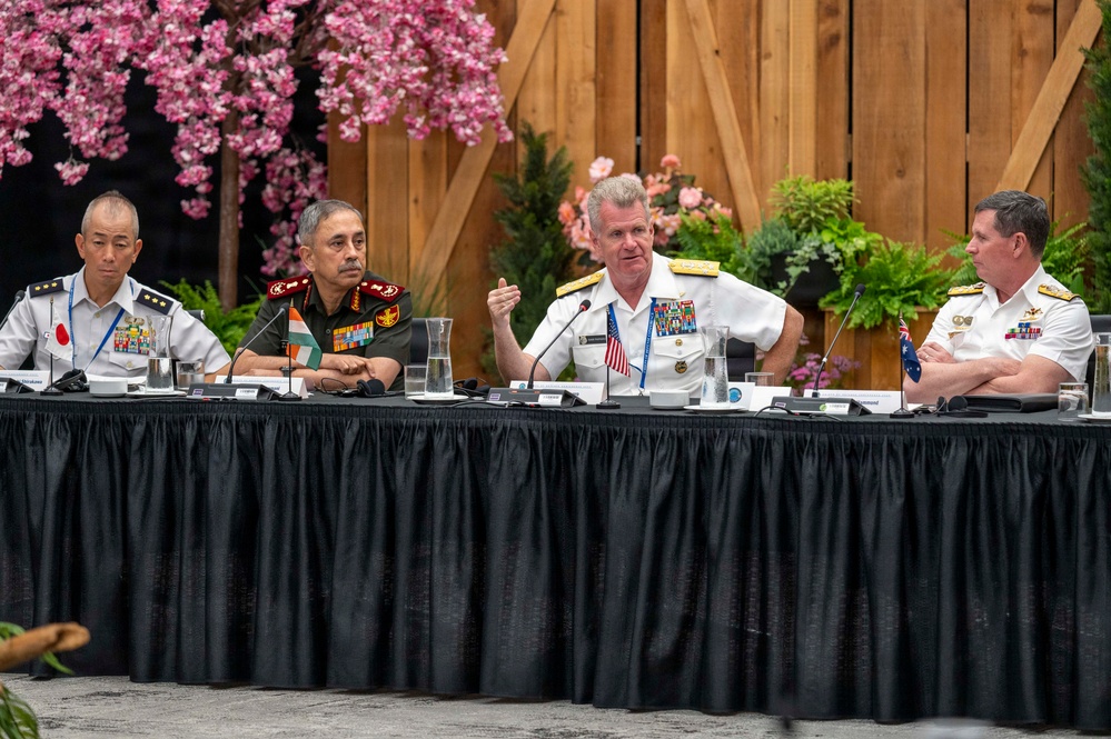 Four senior military officers from different nations are seated at a conference table during what appears to be a multinational military summit or forum. The officers represent at least three identifiable nations based on visible flag pins and uniform styles: Japan, India, the United States, and Australia. The American officer in white naval dress uniform appears to be speaking or presenting at the panel.