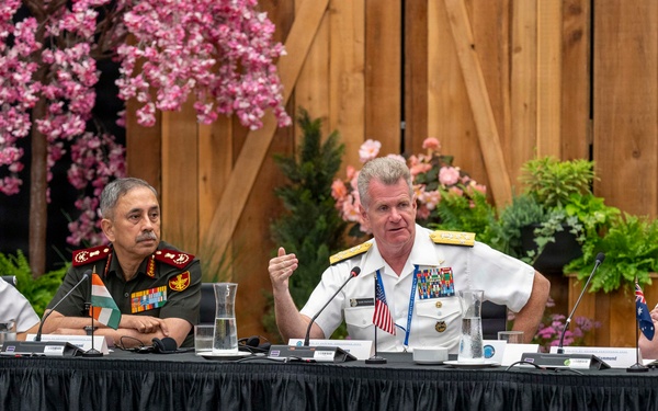 Four senior military officers from different nations are seated at a conference table during what appears to be a multinational military summit or forum. The officers represent at least three identifiable nations based on visible flag pins and uniform styles: Japan, India, the United States, and Australia. The American officer in white naval dress uniform appears to be speaking or presenting at the panel.