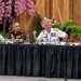 Four senior military officers from different nations are seated at a conference table during what appears to be a multinational military summit or forum. The officers represent at least three identifiable nations based on visible flag pins and uniform styles: Japan, India, the United States, and Australia. The American officer in white naval dress uniform appears to be speaking or presenting at the panel.