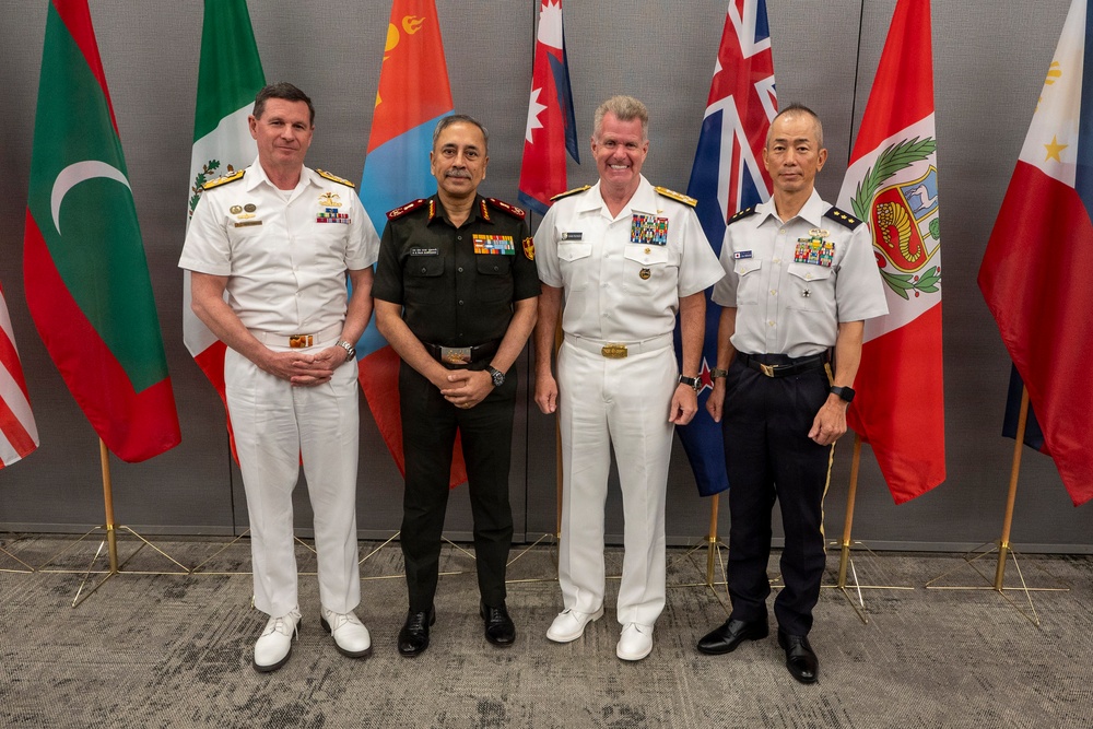 Four senior military officers from different nations pose for a formal group photograph in front of a row of international flags. The officers are wearing distinct uniforms representing naval and army branches from what appear to be multiple allied nations. The setting appears to be an official multilateral military conference or summit.