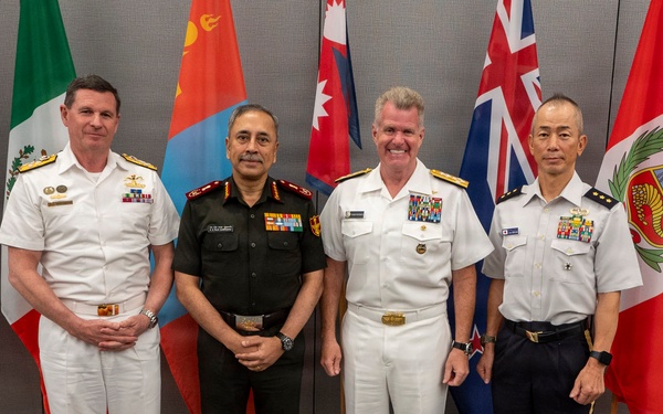 Four senior military officers from different nations pose for a formal group photograph in front of a row of international flags. The officers are wearing distinct uniforms representing naval and army branches from what appear to be multiple allied nations. The setting appears to be an official multilateral military conference or summit.