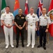Four senior military officers from different nations pose for a formal group photograph in front of a row of international flags. The officers are wearing distinct uniforms representing naval and army branches from what appear to be multiple allied nations. The setting appears to be an official multilateral military conference or summit.