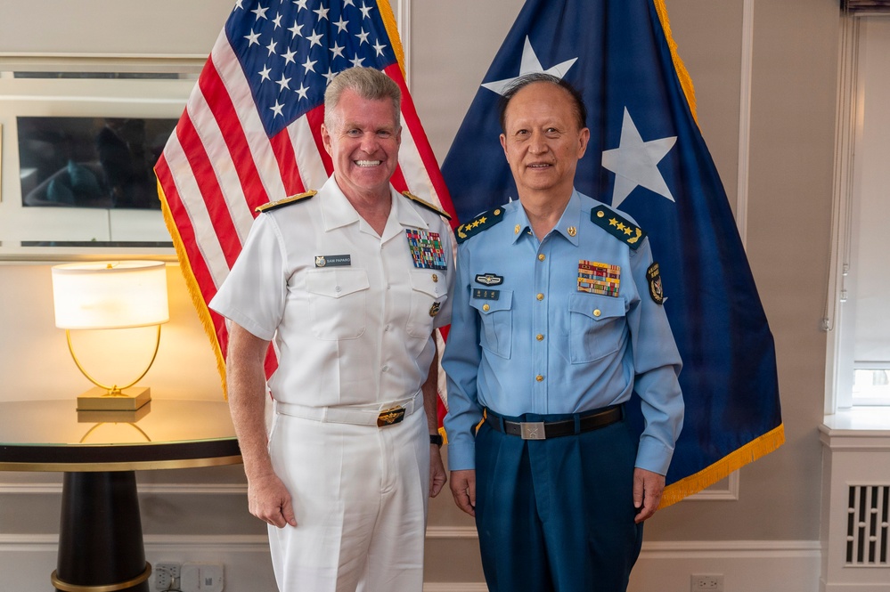 Two senior military officers are posing for a formal photograph in what appears to be an official office setting. One officer is wearing a white U.S. Navy dress uniform with multiple ribbons and insignia, while the other is wearing a light blue military uniform consistent with Chinese People's Liberation Army Air Force (PLAAF) dress attire with four-star rank insignia. Both flags visible in the background suggest this is a bilateral military meeting.