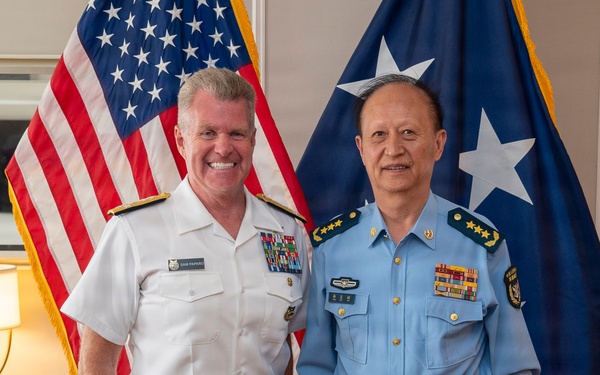 Two senior military officers are posing for a formal photograph in what appears to be an official office setting. One officer is wearing a white U.S. Navy dress uniform with multiple ribbons and insignia, while the other is wearing a light blue military uniform consistent with Chinese People's Liberation Army Air Force (PLAAF) dress attire with four-star rank insignia. Both flags visible in the background suggest this is a bilateral military meeting.