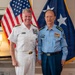 Two senior military officers are posing for a formal photograph in what appears to be an official office setting. One officer is wearing a white U.S. Navy dress uniform with multiple ribbons and insignia, while the other is wearing a light blue military uniform consistent with Chinese People's Liberation Army Air Force (PLAAF) dress attire with four-star rank insignia. Both flags visible in the background suggest this is a bilateral military meeting.