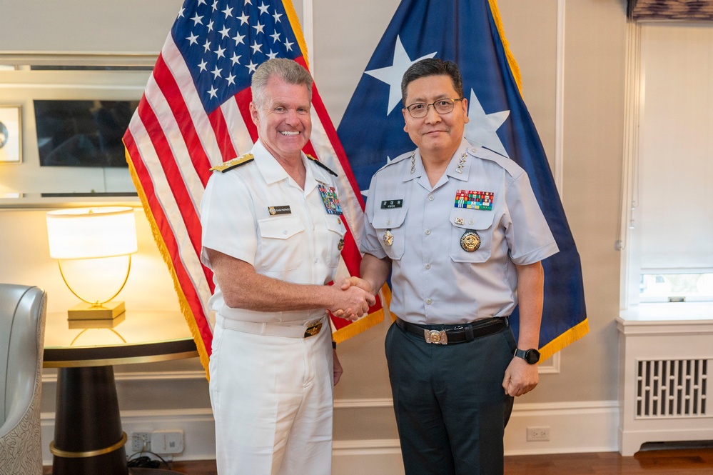 Two military officers in uniform are shaking hands and posing for a formal photograph in what appears to be an official office setting. One officer is wearing a white U.S. Navy dress uniform with admiral rank insignia, while the other is wearing a light gray uniform consistent with a senior Asian military officer. An American flag and a two-star admiral's flag are visible in the background.