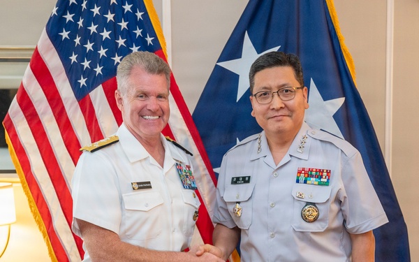 Two military officers in uniform are shaking hands and posing for a formal photograph in what appears to be an official office setting. One officer is wearing a white U.S. Navy dress uniform with admiral rank insignia, while the other is wearing a light gray uniform consistent with a senior Asian military officer. An American flag and a two-star admiral's flag are visible in the background.