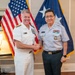 Two military officers in uniform are shaking hands and posing for a formal photograph in what appears to be an official office setting. One officer is wearing a white U.S. Navy dress uniform with admiral rank insignia, while the other is wearing a light gray uniform consistent with a senior Asian military officer. An American flag and a two-star admiral's flag are visible in the background.