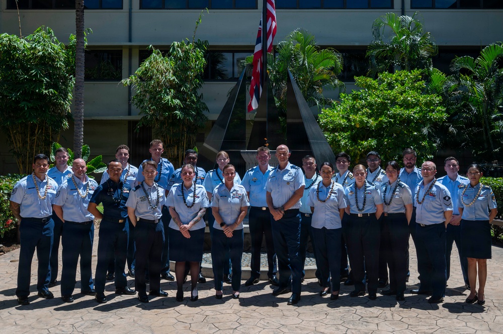A group of approximately 25 military personnel in light blue uniforms are posed for a formal group photograph outdoors. Several individuals are wearing traditional Hawaiian lei garlands, suggesting a ceremonial or welcoming occasion in Hawaii. They are standing in front of a modern sculptural monument with a flagpole displaying what appears to be the Hawaii state flag alongside the US flag.