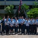 A group of approximately 25 military personnel in light blue uniforms are posed for a formal group photograph outdoors. Several individuals are wearing traditional Hawaiian lei garlands, suggesting a ceremonial or welcoming occasion in Hawaii. They are standing in front of a modern sculptural monument with a flagpole displaying what appears to be the Hawaii state flag alongside the US flag.