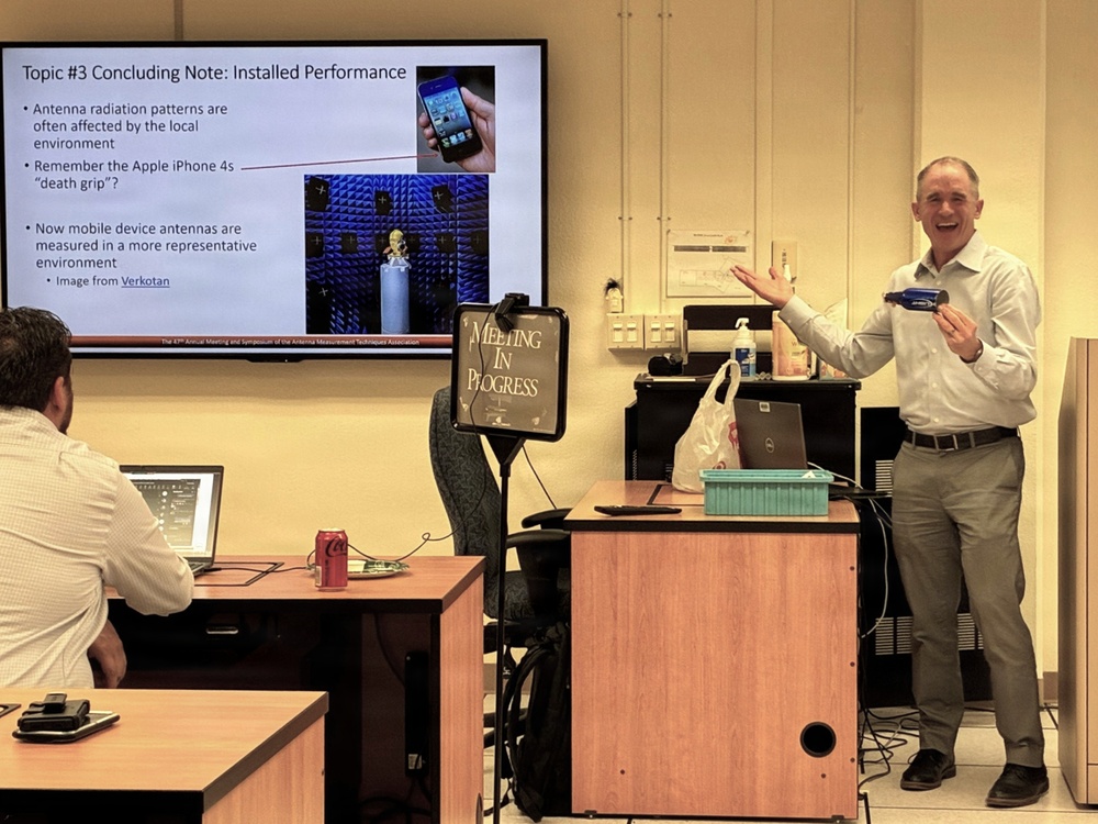 A presenter is delivering a technical lecture on antenna performance at what appears to be the 47th Annual Meeting and Symposium of the Antenna Measurement Techniques Association (AMTA). The slide on the large display screen covers 'Topic #3 Concluding Note: Installed Performance,' referencing the Apple iPhone 4s 'death grip' antenna issue and modern measurement environments. The presenter is standing to the right of the screen, smiling and gesturing enthusiastically while holding a laser pointer or clicker.