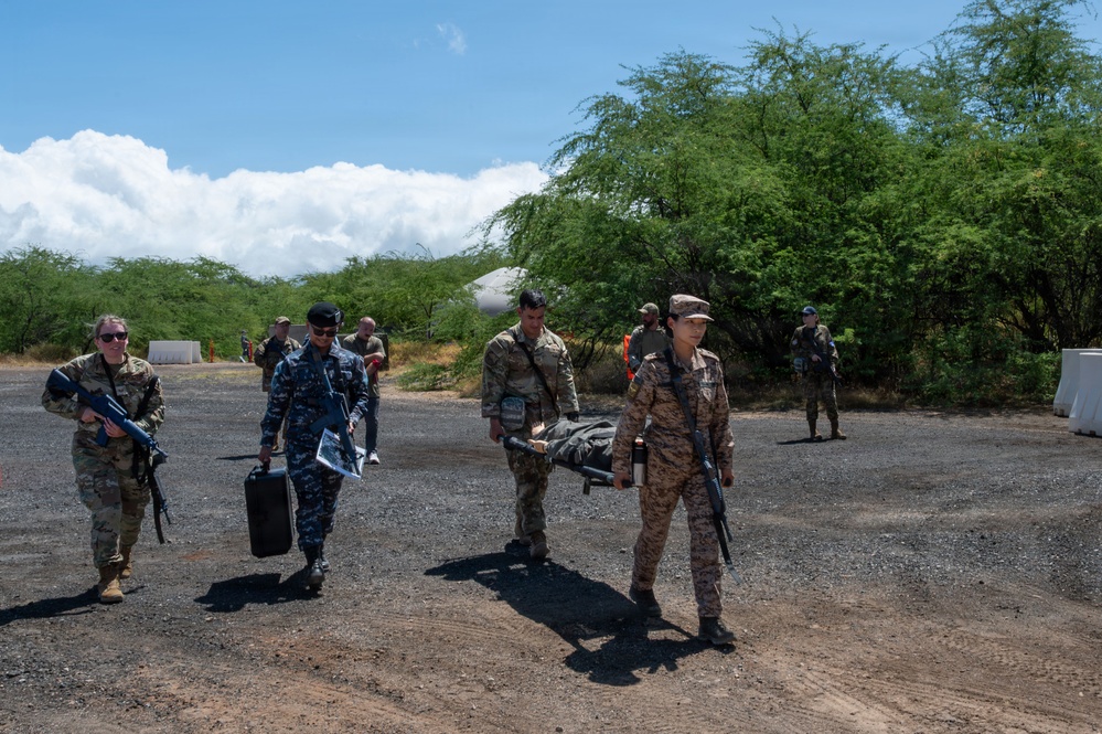 A group of military personnel from multiple nations are conducting what appears to be a joint training exercise in an outdoor area with gravel terrain and tropical vegetation. Several soldiers in different uniforms are carrying a stretcher with a simulated casualty, while others carry weapons and equipment. The scene suggests a multinational military medical or humanitarian assistance drill.