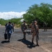 A group of military personnel from multiple nations are conducting what appears to be a joint training exercise in an outdoor area with gravel terrain and tropical vegetation. Several soldiers in different uniforms are carrying a stretcher with a simulated casualty, while others carry weapons and equipment. The scene suggests a multinational military medical or humanitarian assistance drill.