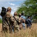 A group of five military personnel in various camouflage uniforms are walking in a single-file line through tall dry grass toward a treeline. At least two distinct camouflage patterns are visible, suggesting a multinational or joint-service exercise. The group is moving away from the camera in an outdoor field environment with scattered trees and a partly cloudy sky.