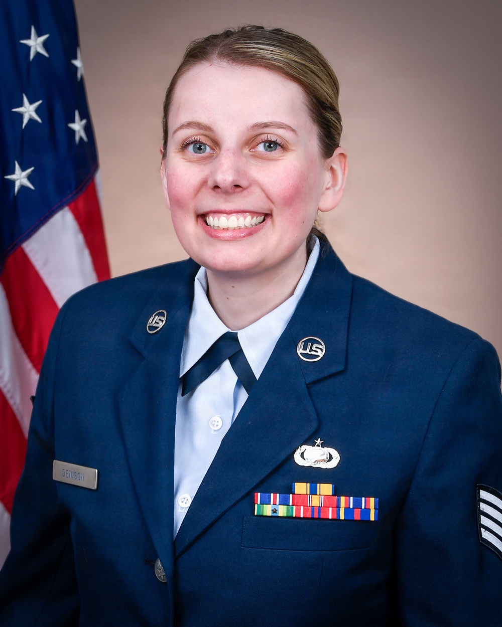 A formal military portrait of a U.S. Air Force enlisted member in dress blue uniform, smiling in front of an American flag. The subject wears the rank of Staff Sergeant (visible chevrons on sleeve) and displays multiple service ribbons and a Senior badge with star device. The name tape reads 'DENISON'.