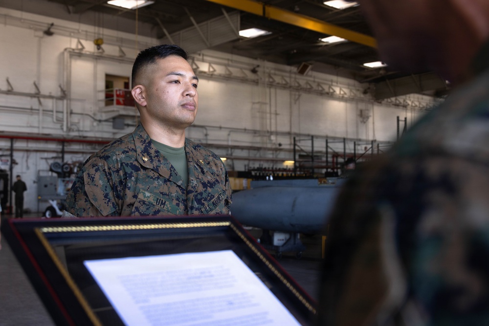 A U.S. Marine in woodland MARPAT camouflage uniform stands at attention inside what appears to be a military hangar or maintenance facility. A framed document or certificate is being presented to the Marine, suggesting a formal recognition or award ceremony. The setting includes visible military equipment and infrastructure in the background.
