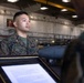 A U.S. Marine in woodland MARPAT camouflage uniform stands at attention inside what appears to be a military hangar or maintenance facility. A framed document or certificate is being presented to the Marine, suggesting a formal recognition or award ceremony. The setting includes visible military equipment and infrastructure in the background.