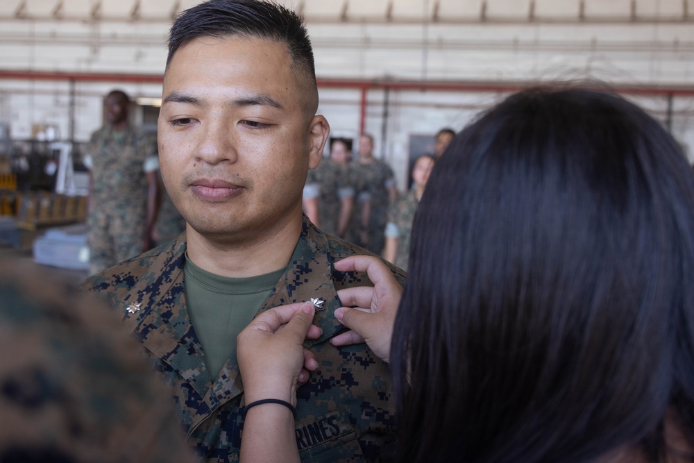 A U.S. Marine in MARPAT camouflage uniform is having rank insignia pinned onto his collar during what appears to be a promotion ceremony. A person with dark hair is pinning a silver oak leaf insignia (Major rank) onto the right collar of the Marine's uniform. The ceremony is taking place inside a military hangar with other uniformed personnel visible in the background.