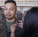 A U.S. Marine in MARPAT camouflage uniform is having rank insignia pinned onto his collar during what appears to be a promotion ceremony. A person with dark hair is pinning a silver oak leaf insignia (Major rank) onto the right collar of the Marine's uniform. The ceremony is taking place inside a military hangar with other uniformed personnel visible in the background.
