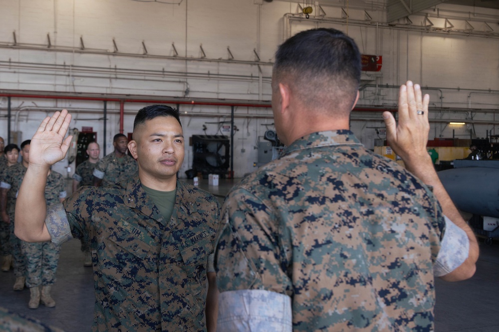 Two U.S. Marines in MARPAT camouflage uniforms are conducting what appears to be a reenlistment or oath of office ceremony inside a military hangar. Both individuals have their right hands raised, with one Marine facing the camera and the other with his back to it. Additional Marines are visible standing in formation in the background.