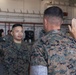 Two U.S. Marines in MARPAT camouflage uniforms are conducting what appears to be a reenlistment or oath of office ceremony inside a military hangar. Both individuals have their right hands raised, with one Marine facing the camera and the other with his back to it. Additional Marines are visible standing in formation in the background.