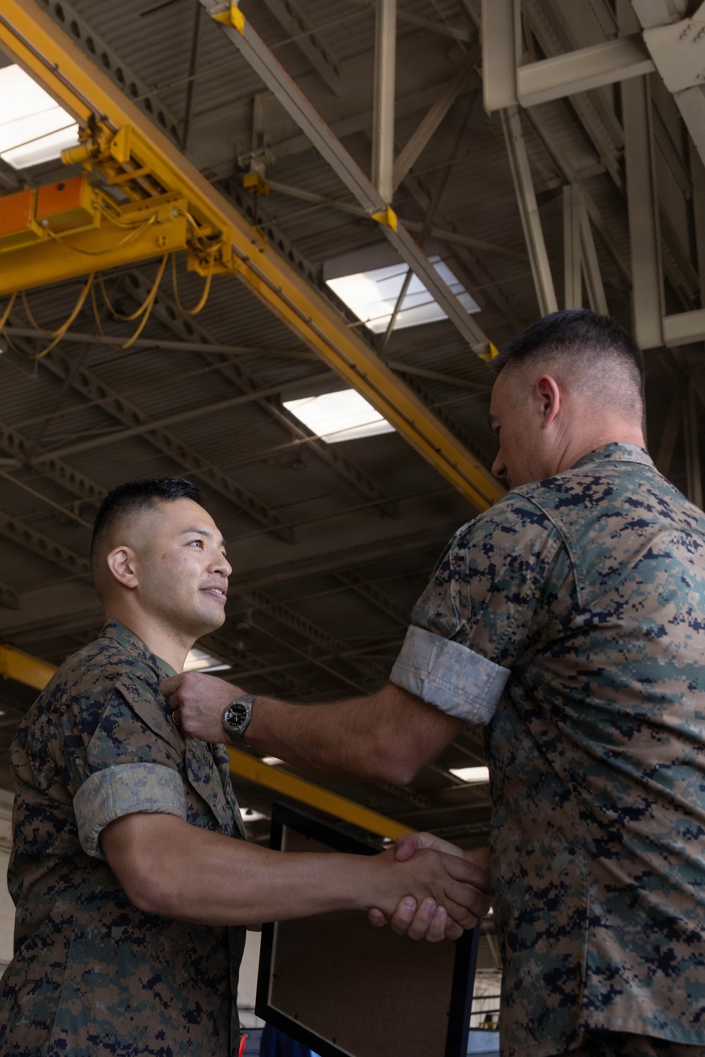Two U.S. Marines in MARPAT digital camouflage uniforms are engaged in what appears to be a military awards or promotion ceremony inside an industrial hangar facility. One Marine is pinning something onto the chest of the other while simultaneously shaking his hand, a common practice during military recognition ceremonies. The setting features overhead yellow crane infrastructure typical of an aircraft maintenance hangar.