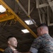 Two U.S. Marines in MARPAT digital camouflage uniforms are engaged in what appears to be a military awards or promotion ceremony inside an industrial hangar facility. One Marine is pinning something onto the chest of the other while simultaneously shaking his hand, a common practice during military recognition ceremonies. The setting features overhead yellow crane infrastructure typical of an aircraft maintenance hangar.