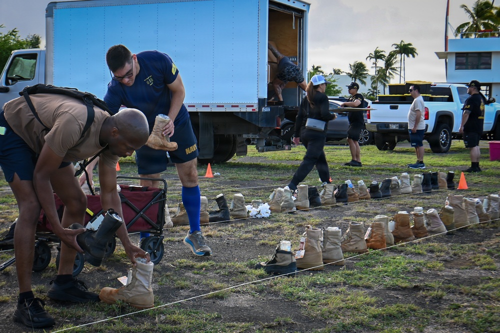 Military personnel and civilians are organizing rows of combat boots on a grassy outdoor area, likely as part of a memorial display or tribute event. Two individuals in the foreground are carefully placing boots along a rope line, while others work in the background near a large box truck. The tropical setting with palm trees and the mix of Navy uniforms suggests this may be taking place at a military installation in Hawaii.