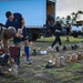 Military personnel and civilians are organizing rows of combat boots on a grassy outdoor area, likely as part of a memorial display or tribute event. Two individuals in the foreground are carefully placing boots along a rope line, while others work in the background near a large box truck. The tropical setting with palm trees and the mix of Navy uniforms suggests this may be taking place at a military installation in Hawaii.