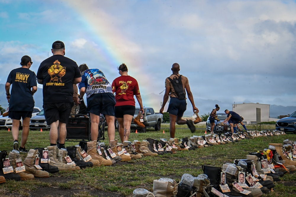 A group of people walk alongside rows of military boots arranged on a grass field, each pair accompanied by a photo card, representing fallen service members in a memorial display. A rainbow is visible in the background sky, adding a poignant visual element to the solemn occasion. The setting appears to be a military installation or base, likely in Hawaii based on visible shirt text.
