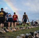 A group of people walk alongside rows of military boots arranged on a grass field, each pair accompanied by a photo card, representing fallen service members in a memorial display. A rainbow is visible in the background sky, adding a poignant visual element to the solemn occasion. The setting appears to be a military installation or base, likely in Hawaii based on visible shirt text.