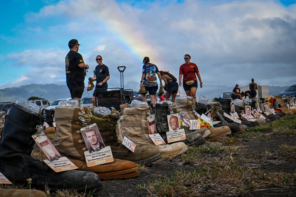 A memorial display featuring military combat boots lined up on the ground, each tagged with identification cards bearing photos and names of fallen service members. The boots appear to represent soldiers killed in Operation Enduring Freedom and Operation Iraqi Freedom, with visible tags including 'Army SPC Robert J Cook' who died 01/29/2004. A rainbow is visible in the background sky above a group of people who appear to be participating in or organizing the memorial event.