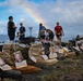 A memorial display featuring military combat boots lined up on the ground, each tagged with identification cards bearing photos and names of fallen service members. The boots appear to represent soldiers killed in Operation Enduring Freedom and Operation Iraqi Freedom, with visible tags including 'Army SPC Robert J Cook' who died 01/29/2004. A rainbow is visible in the background sky above a group of people who appear to be participating in or organizing the memorial event.