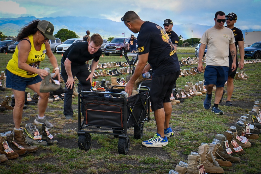 A group of people are setting up or arranging a memorial display consisting of hundreds of military boots placed across a grassy field, each accompanied by what appear to be photographs and name tags. The event appears to be a 'Boot Memorial,' as indicated by the yellow shirt worn by one participant. The setting appears to be an outdoor field in Hawaii, suggested by the mountainous backdrop and tropical vegetation.