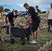 A group of people are setting up or arranging a memorial display consisting of hundreds of military boots placed across a grassy field, each accompanied by what appear to be photographs and name tags. The event appears to be a 'Boot Memorial,' as indicated by the yellow shirt worn by one participant. The setting appears to be an outdoor field in Hawaii, suggested by the mountainous backdrop and tropical vegetation.