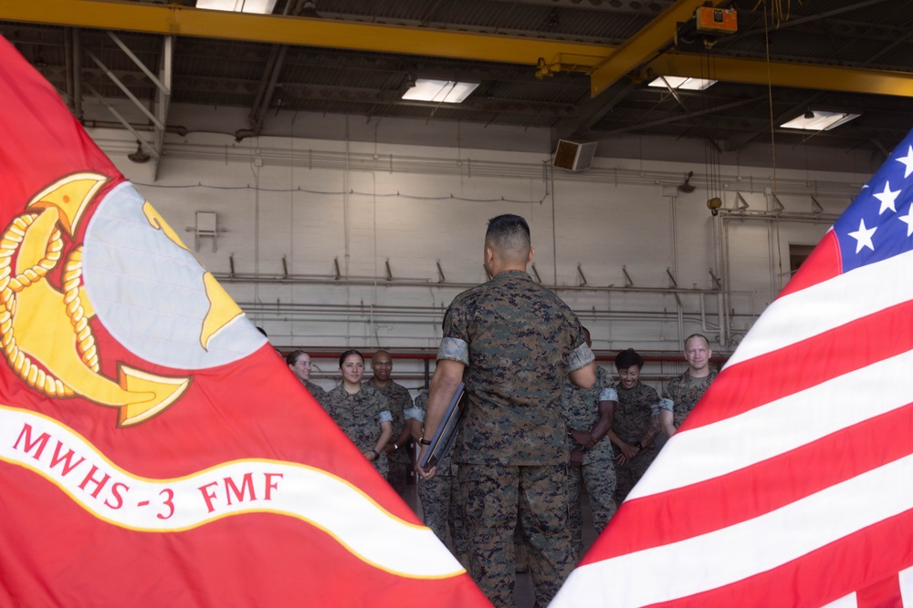A military formation or briefing is taking place inside what appears to be a hangar facility. A Marine in camouflage utilities stands with his back to the camera, addressing a group of fellow Marines. Two flags are prominently visible in the foreground: the United States flag and a unit flag for MWHS-3 FMF (Marine Wing Headquarters Squadron 3, Fleet Marine Force).