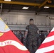 A military formation or briefing is taking place inside what appears to be a hangar facility. A Marine in camouflage utilities stands with his back to the camera, addressing a group of fellow Marines. Two flags are prominently visible in the foreground: the United States flag and a unit flag for MWHS-3 FMF (Marine Wing Headquarters Squadron 3, Fleet Marine Force).