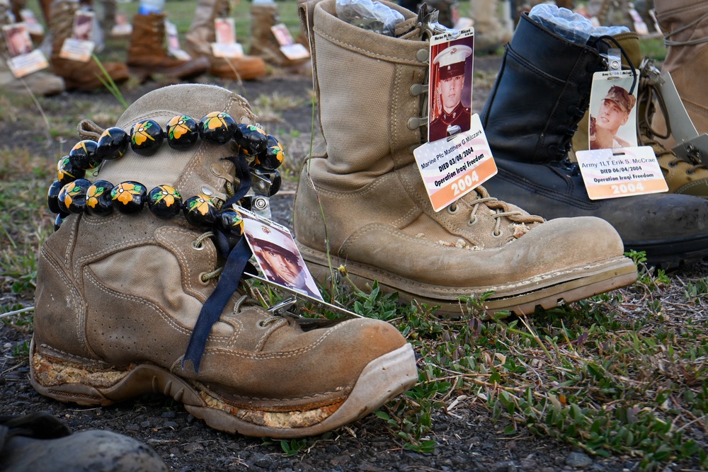 This image depicts a memorial display featuring military combat boots arranged on the ground, each bearing identification tags with photographs and information about fallen service members from Operation Iraqi Freedom. The boots represent soldiers and Marines who died in 2004, serving as a tribute installation. One boot in the foreground is adorned with a decorative beaded lei, adding a personal touch to the memorial.