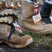 This image depicts a memorial display featuring military combat boots arranged on the ground, each bearing identification tags with photographs and information about fallen service members from Operation Iraqi Freedom. The boots represent soldiers and Marines who died in 2004, serving as a tribute installation. One boot in the foreground is adorned with a decorative beaded lei, adding a personal touch to the memorial.
