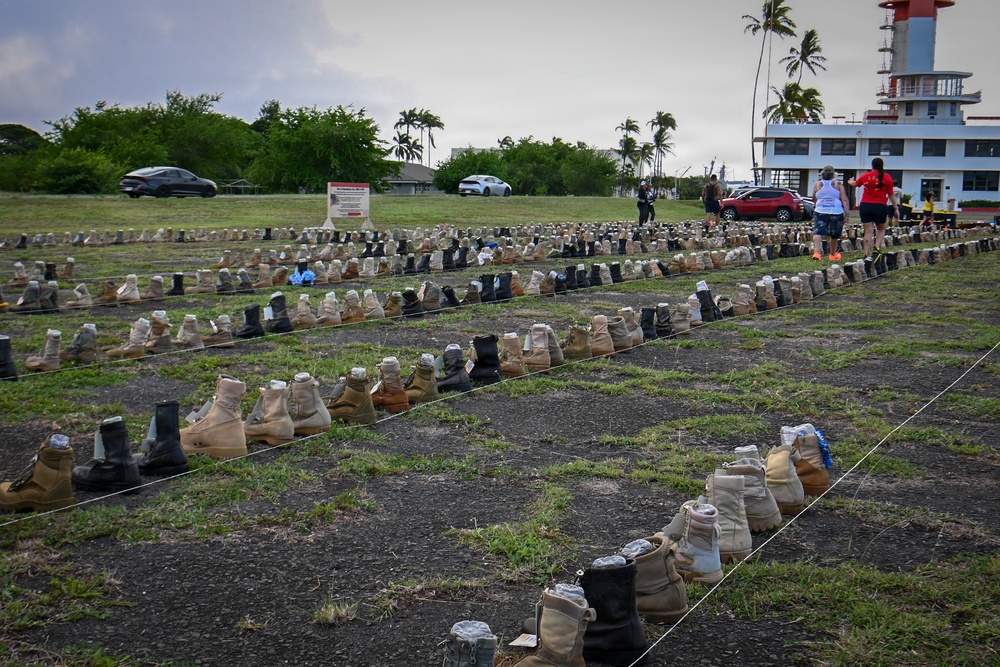 A large outdoor memorial display featuring hundreds of military combat boots arranged in rows on a grassy field, likely representing fallen or deceased veterans. The installation appears to be a tribute or awareness display, possibly related to veteran suicide or combat casualties. The setting appears to be a military installation or public grounds in a tropical location, evidenced by palm trees and a distinctive tower structure in the background.