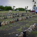 A large outdoor memorial display featuring hundreds of military combat boots arranged in rows on a grassy field, likely representing fallen or deceased veterans. The installation appears to be a tribute or awareness display, possibly related to veteran suicide or combat casualties. The setting appears to be a military installation or public grounds in a tropical location, evidenced by palm trees and a distinctive tower structure in the background.