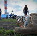 A row of worn military combat boots is arranged on grass in the foreground, each containing small tags or notes, suggesting a memorial or tribute display. Several people are visible in the background walking and gathering near a red-and-white striped tower structure, likely a lighthouse or navigational beacon. The scene appears to be an outdoor commemorative event, possibly honoring fallen service members.